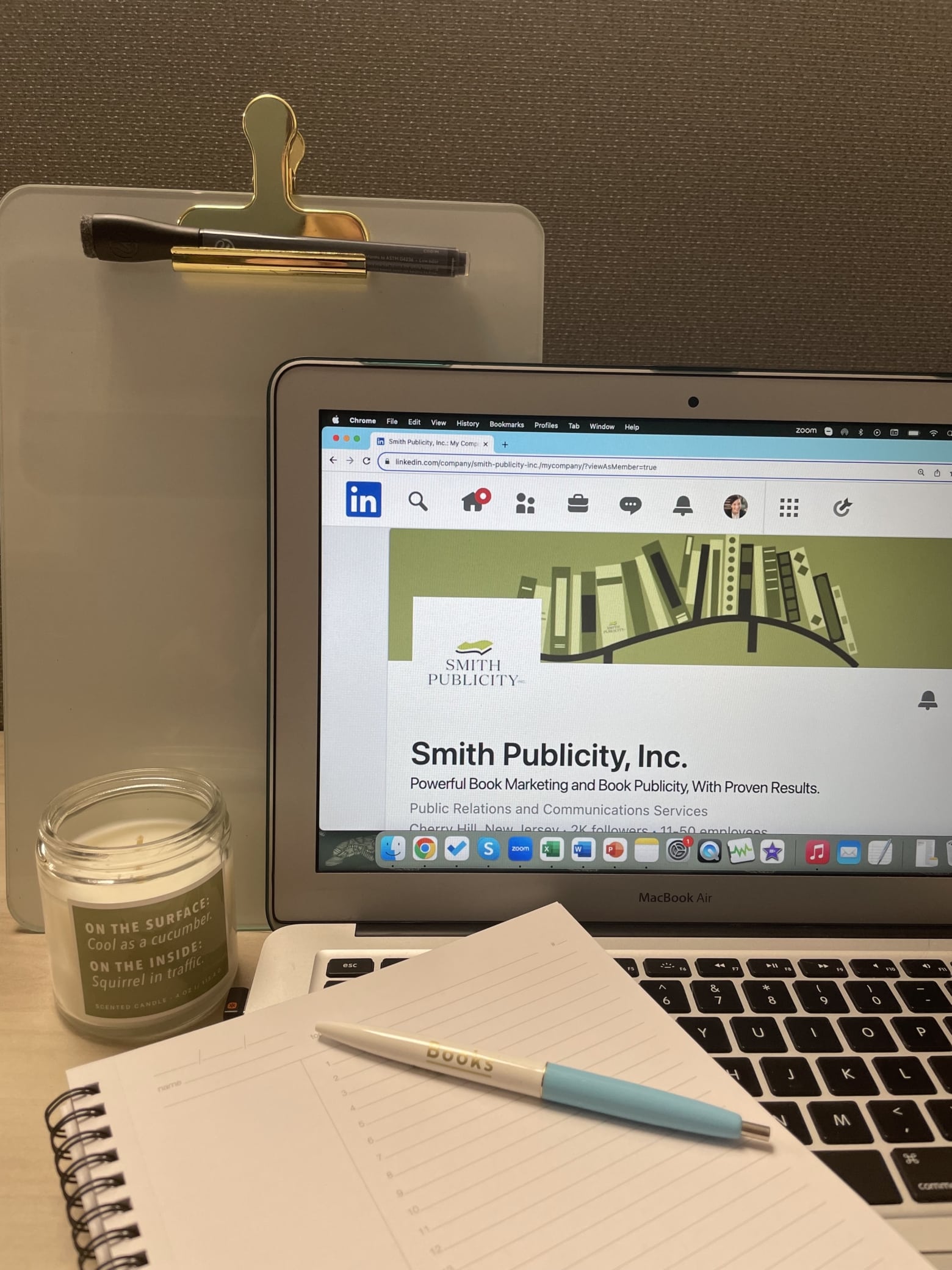 A notepad and pen are placed alongside a laptop, opened to Smith Publicity's LinkedIn page. There is a matching green candle next to the laptop.