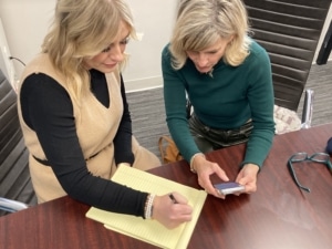 Publicity Manager Courtney Link takes notes on a yellow legal pad with a pen on book marketing strategy while VP of Publicity Janet Shapiro references something on her phone.